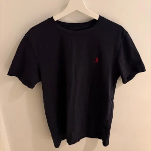 Svart t-shirt från Polo Ralph Lauren XL - Klassisk svart t-shirt från Polo Ralph Lauren i storlek XL. T-shirten har korta ärmar, rund halsringning och en liten röd broderad logga på bröstet. Tillverkad i mjuk bomull för en skön känsla och stilren look. Passar de som har L