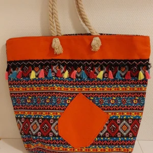 Färgglad väska med tofsar - Nyskick. Stor och färgglad väska i bomull med orange ovandel, mönstrad botten i blått, rött och gult samt dekorativa tofsar i flera färger. Väskan har grova rep-handtag och en orange ficka framtill. Perfekt för att bära allt du behöver. 