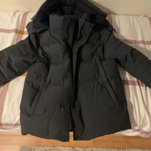 Svart dunjacka från Canada Goose - Säljer en svart dunjacka från Canada Goose med huva och dragkedja framtill. Jackan har en klassisk pufferstil, diskret logga på ärmen och är perfekt för kalla dagar. Fylld med dun och fjädrar för extra värme och har praktiska fickor på sidorna.