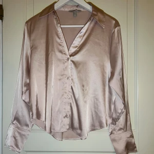 Champagnefärgad satinskjorta - Snygg champagnefärgad skjorta i satin med klassisk krage och knappar framtill. Skjortan har långa ärmar med breda manschetter och en lätt glansig finish som ger en lyxig känsla. Perfekt för dig som vill sticka ut med en elegant look. Den finns fläckar på skjortan.