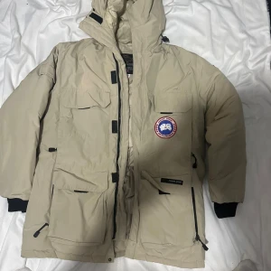 Beige dunjacka från Canada Goose L - Säljer en beige dunjacka från Canada Goose i storlek L. Jackan har flera stora fickor, huva och klassisk logga på bröstet. Perfekt för kalla dagar med sin tjocka vaddering och quiltad insida. Stängs med dragkedja och kardborre. Snygg och funktionell vinterjacka.