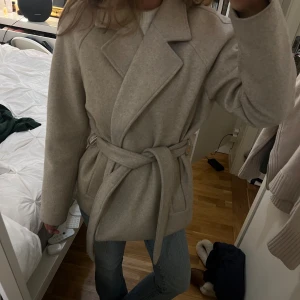 Beige kappa med bälte och krage - Snygg beige kappa med bred krage och knytskärp i midjan. Jackan har en klassisk, rak passform och är tillverkad i ett mjukt ull-liknande material. Perfekt för dig som gillar stilrena och tidlösa plagg.