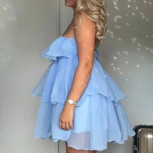 Ljusblå volangklänning axelbandslös - Superfin ljusblå axelbandslös klänning med flera lager volanger som ger en fluffig och drömmig look. Klänningen är kort och har en luftig känsla, perfekt för varma dagar. Materialet är tunt och lätt, troligtvis polyester eller liknande.