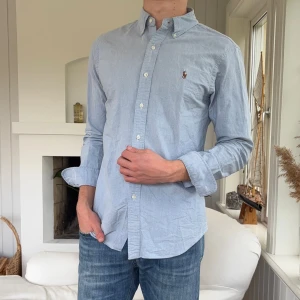 Ljusblå skjorta Polo Ralph Lauren - Klassisk ljusblå skjorta från Polo Ralph Lauren i slim fit-modell. Skjortan har button down-krage, broderad logga på bröstet och vita knappar. Tillverkad i bomull och har långa ärmar. Perfekt för en clean och stilren look.