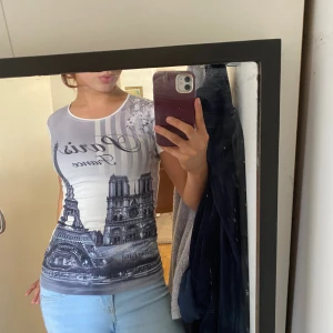 Vit och grå Paris-mönstrad topp - Säljer en tight topp med tryck av Eiffeltornet och Notre Dame i svartvitt. Toppen är ärmlös och har rund halsringning. Texten 'Paris France' syns framtill. Perfekt för dig som gillar unika motiv och vill sticka ut.