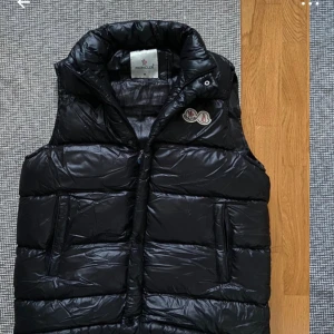 Svart dunväst från Moncler med huva - Svart glansig dunväst från Moncler med avtagbar huva och klassisk logga på bröstet. Västen har dragkedja framtill, två fickor med dragkedja och en quiltad design som ger en riktigt clean look. Perfekt för lager-på-lager under kyliga dagar.