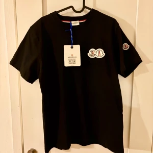 Svart Moncler t-shirt med logga - Svart t-shirt från Moncler i bomull med klassisk passform. T-shirten har korta ärmar och snygga loggpatchar på bröstet och ena ärmen. Perfekt för dig som gillar stilrena och exklusiva plagg med tydlig branding.