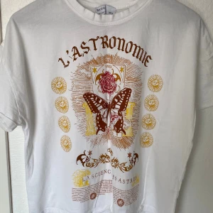 Vit t-shirt med tryck från Bershka - Vit t-shirt från Bershka med coolt tryck i brunt och gult framtill. Motivet har texten 'L'ASTRONOMIE', fjäril, ros och solsymboler. Klassisk rund hals och korta ärmar. Skön och mjuk i bomull, perfekt för dig som gillar unika prints.