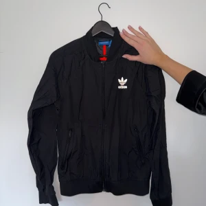 Svart vindjacka från Adidas - Svart vindjacka från Adidas med klassisk vit logga på bröstet. Jackan har dragkedja framtill, två fickor med dragkedja och ribbade muddar vid ärmslut och nederkant. Perfekt för en sportig och stilren look.