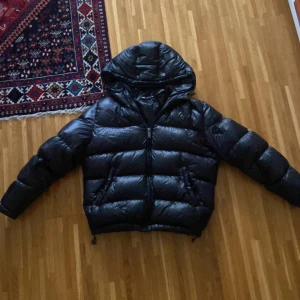 Svart pufferjacka med huva - Säljer en svart pufferjacka med glansig finish och stor huva. Jackan har dragkedja framtill och breda, vadderade paneler som ger en riktigt fet look. Perfekt för kalla dagar när du vill hålla stilen och värmen.