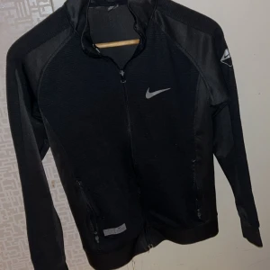 Svart träningsjacka från Nike - Svart träningsjacka från Nike med hel dragkedja framtill och två fickor med dragkedja. Jackan har lång ärm, sportig look och diskret Nike-logga på bröstet och ärmen. Perfekt för dig som gillar en clean och stilren stil.         Den är liten i storleken. 