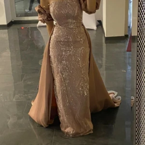 Champagnefärgad glittrig långklänning - Säljer en elegant ljus rosa långklänning med glittriga detaljer och figurnära passform. Klänningen har avtagbara puffärmar och en snygg slits på sidan. Perfekt för bal eller bröllop tillfällen. Materialet känns lyxigt och skimrande. Den är skräddarsydd i Turkiet och har använt den bara under några timmar. Den är storlek S men hade passat en M också 