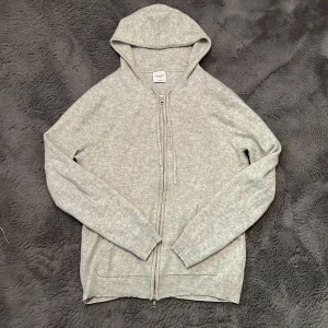 Grå kashmirhoodie från John Henric - Mjuk och lyxig grå hoodie i 100% kashmir från John Henric. Tröjan har dragkedja framtill, snörning i huvan och ribbade muddar. Perfekt för dig som vill ha en stilren och bekväm look med premiumkänsla.