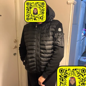 Svart pufferjacka från Moncler - Säljer en svart pufferjacka från Moncler med huva och dragkedja. Jackan har quiltad framsida i glansigt material, stickade ärmar och en snygg ficka med dragkedja på ena ärmen. Klassisk Moncler-logga på axeln. Perfekt för kalla dagar.