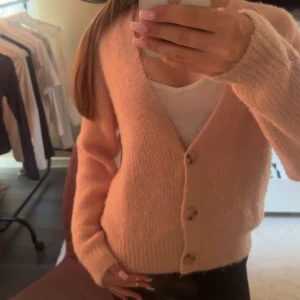 Ljusrosa kofta med knappar - Supermysig ljusrosa kofta med v-ringning och tre knappar framtill. Koftan har lång ärm och är stickad i ett fluffigt material som känns riktigt skönt mot huden. Perfekt att slänga på sig när det är lite kyligare ute.