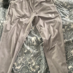 Grå mjukisbyxor från Under Armour - Snygga grå mjukisbyxor från Under Armour med dragsko i midjan och logga på låret. Byxorna har en avslappnad passform och är perfekta för chill eller träning. Tillverkade i mjukt och skönt material.