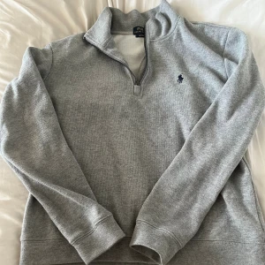 Grå half zip tröja från Polo Ralph Lauren - Snygg grå långärmad tröja från Polo Ralph Lauren med half zip och klassisk broderad logga på bröstet. Tröjan har en hög krage och är tillverkad i mjuk bomullsmix, perfekt för lager-på-lager under höst och vinter.