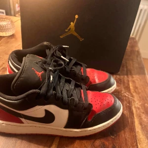 Nike Air Jordan 1 Low  - Säljer ett par Nike Air Jordan 1 Low i den klassiska Bred-färgställningen. Skorna har svart, rött och vitt skinn, svart swoosh och snörning. Ikonisk Air Jordan-logga på hälen och Jumpman på plösen. Perfekta för dig som gillar streetwear och sneakers med attityd.