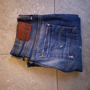 Blå G-Star RAW jeans straight fit - Säljer ett par klassiska blå jeans från G-Star RAW med snygga slitningar och raka ben. Jeansen har fem fickor, tydliga kontrastsömmar och en stor brun logopatch bak. Perfekta för dig som gillar en tidlös och avslappnad stil.
