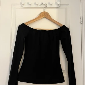 Svart offshoulder topp - En stilren svart offshoulder topp med långa ärmar. Toppen har en enkel och smickrande passform som framhäver axlarna. Perfekt att matcha med både jeans och kjol för en trendig look. Endast testad. 