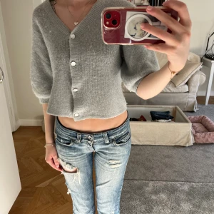 Ljusgrå croppad kofta med knappar - Superfin ljusgrå kofta med croppad passform och stora vita knappar framtill. Tröjan har trekvartsärmar och är stickad i ett mjukt material som känns skönt mot huden. Perfekt att slänga på sig över ett linne eller t-shirt.