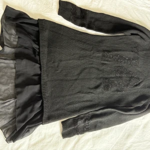 Svart stickad klänning Sisley  - Svart stickad klänning från Sisley med glittriga detaljer på ärmar och framsida. Nederdelen har ett lager av tunnare, transparent tyg som ger en snygg volang-effekt. Långärmad och perfekt för dig som gillar en cool och stilren look. 150 cm