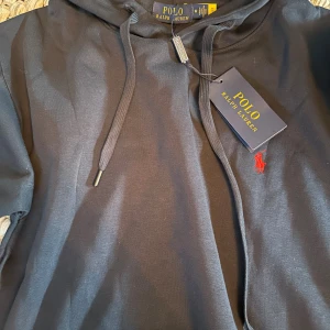 Svart hoodie från Polo Ralph Lauren i storlek m men sitter mer som en S - Svart hoodie från Polo Ralph Lauren med klassisk huva, snörning och känguruficka. Tröjan har en röd broderad logga på bröstet och är tillverkad i mjuk bomull. Perfekt för en chill och stilren look.