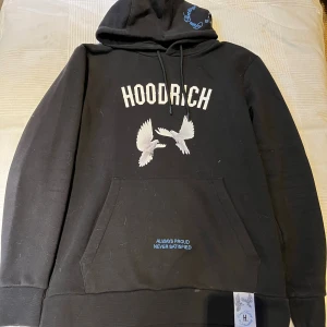 Svart hoodie från Hoodrich med tryck  - Svart hoodie från Hoodrich med vit text och motiv av duvor på både fram- och baksida. Stora tryck med texten 'HOODRICH' och 'From Nothing To Something'. Huva med snörning och känguruficka framtill. Snygga blå detaljer och broderad text. passar som S
