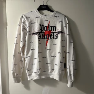 Vit Palm Angels sweatshirt med tryck - Vit sweatshirt från Palm Angels (EJ ORIGINAL) med svart logotyp- och textprint över hela tröjan samt en stor röd blixt och taggtrådsmotiv framtill. Rund halsringning, ribbade muddar och långärmad. Perfekt statement piece för dig som gillar streetwear.