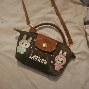 Brun axelväska med Labubu-tryck - Säljer en brun axelväska med gulligt Labubu-tryck i pastellfärger. Väskan har ett handtag och en justerbar axelrem i brunt skinn, samt lock med guldfärgad knapp. Perfekt storlek för det viktigaste och har en unik, lekfull design.