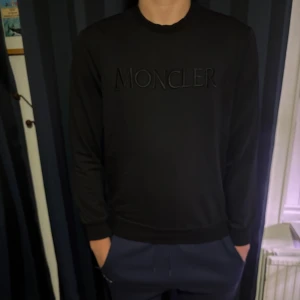 Svart Moncler sweatshirt i bomull - Svart sweatshirt från Moncler med broderad logga på bröstet och emblem på ärmen. Tröjan har rund halsringning och ribbade muddar. Tillverkad i mjuk bomull, perfekt för en clean och stilren look.
