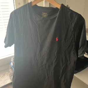 Svart t-shirt från Polo Ralph Lauren - Svart t-shirt från Polo Ralph Lauren med klassisk passform och korta ärmar. Ikoniska röda Polo-loggan broderad på bröstet. Tillverkad i mjuk bomull och har rund halsringning. Perfekt basic-plagg med stilren look.