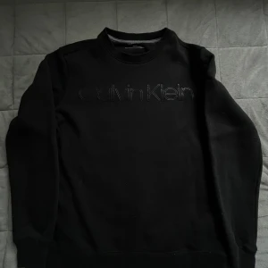 Svart sweatshirt från Calvin Klein XS - Svart sweatshirt från Calvin Klein i klassisk modell med broderad logga framtill. Tröjan har rund halsringning, långa ärmar och ribbade muddar. Perfekt för en clean och stilren look.