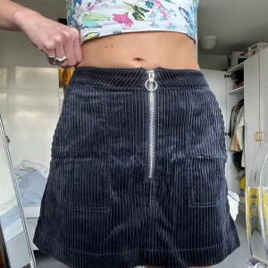 Svart manchesterkjol från Weekday - Supersnygg svart kortkjol i manchester från Weekday med markerade lodräta ränder och en cool silverfärgad dragkedja framtill med ringdetalj. Kjolen har två fickor fram och sitter tajt i midjan. Perfekt att styla med crop top eller stickad tröja.