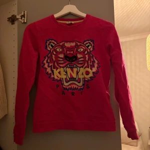 Rosa Kenzo tröja med tigertryck - Säljer en rosa långärmad tröja från Kenzo i storlek XS. Tröjan har ett stort broderat tigertryck och Kenzo Paris-logga framtill. Mjuk och skön bomullskvalitet, perfekt för dig som gillar statement-plagg och coola prints.