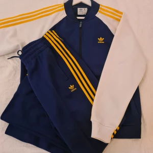 Adidas blå och vit bomberjacka med tillhörande byxa  - Säljer en klassisk Adidas bomberjacka i marinblått och vitt med gula ränder längs ärmarna. Jackan har dragkedja framtill, ribbade muddar och Adidas logga broderad på bröstet. Perfekt för dig som gillar sportig och retro stil. Byxorna storlek medium.