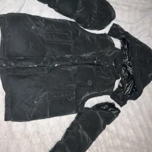 Svart dunväst från Moncler - Svart dunväst från Moncler med quiltad design och diskret logotyp på bröstet. Västen har dragkedja och tryckknappar framtill samt praktiska fickor. Materialet är glansigt och känns lyxigt, perfekt för att hålla värmen under kyliga dagar.