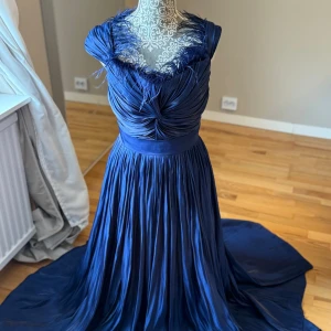 Marinblå långklänning med fjädrar - Elegant marinblå långklänning i glansigt material med plisserad kjol och vackert draperad överdel. Klänningen har fjäderdetaljer vid urringningen, bred midja och knytband i ryggen. Perfekt för dig som vill sticka ut med en lyxig och unik look.