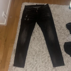 Svarta jeans från Dsquared2, strl 33 - Säljer ett par svarta jeans från Dsquared2 med coola slitningar och tvättade detaljer. Byxorna har klassisk femficksdesign, rakt snitt och patch med 'CIAO' bak i midjan. Tillverkade i Italien och i skönt jeanstyg. Perfekta för dig som gillar streetwear med edge.