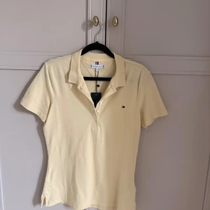 Gul piké från Tommy Hilfiger - Snygg ljusgul piké från Tommy Hilfiger med klassisk krage och korta ärmar. Toppen har knappar framtill och en liten broderad logga på bröstet. 