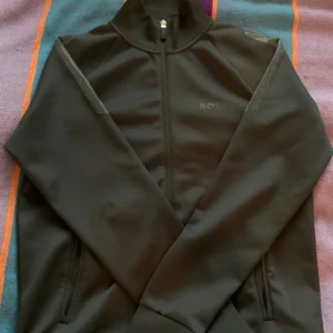 Svart hoddie från BOSS - Svart vindjacka från BOSS med diskret logga på bröstet och hel dragkedja framtill. Jackan har högt krage, långa ärmar och två sidofickor. Materialet är slätt och känns sportigt, perfekt för dig som gillar stilren och enkel design.