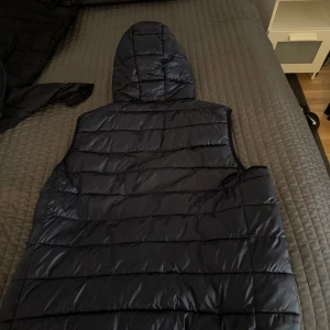 Mörkblå CK dunväst med huva och dragkedja - Snygg svart dunväst med huva och dragkedja framtill. Västen har quiltad design med horisontella sömmar och två fickor med dragkedja. Perfekt för lager-på-lager och enkel att matcha med olika outfits.