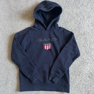 Marinblå hoodie från Gant - Snygg marinblå hoodie från GANT med broderad logga och sköld på bröstet. Klassisk känguruficka framtill, huva och mjukt material i bomull och polyester. Perfekt för dig som gillar stilren och sportig look.