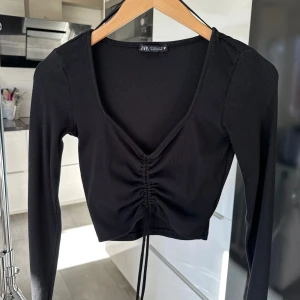 Svart croppad topp med snörning från Zara - Säljer en svart croppad topp från Zara med lång ärm och djup v-ringning. Toppen har ribbad struktur och snygg snörning framtill som ger en rynkad effekt/justera hur lång/kort du vill ha den🖤 storlek M men S passar nog också