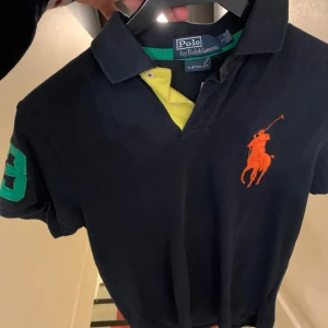 Mörkblå pikétröja Polo Ralph Lauren - Snygg mörkblå pikétröja från Polo Ralph Lauren i bomull. Tröjan har korta ärmar, klassisk krage med gul insida och gröna detaljer. Stor orange logga på bröstet och en grön siffra på ärmen. Perfekt för dig som gillar sportig och stilren look.