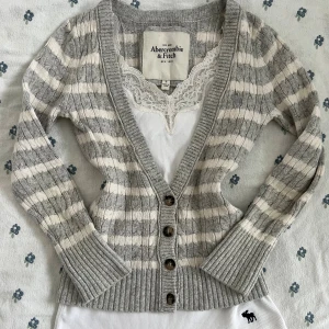 Randig kofta från Abercrombie & Fitch - Supermysig kofta i grått och vitt från Abercrombie & Fitch. Den har kabelstickat mönster, ribbade muddar och knappar framtill. Materialmix med bomull, viskos, akryl och kaninull. Perfekt för lager-på-lager och chill vibes. Spetlinne ingår ej. Det finns inga bilder med den på eftersom den är för liten för mig det är ju varför jag säljer den. Fråga om måtten vid behöv.
