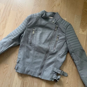 Grå skinnjacka från Voyelles, strl 38 - Snygg grå skinnjacka från Voyelles med coola detaljer som quiltade axlar och ärmar, dragkedjor framtill och på ärmarna samt dubbla bälten i midjan. Jackan har en asymmetrisk dragkedja och ståkrage med tryckknapp. Perfekt för dig som vill ha en edgy look.
