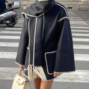 Mörkblå kappa med fransar och huva - Snygg mörkblå kappa med kontrasterande vita sömmar, stor huva och dekorativa fransar nedtill. Jackan har vida ärmar och stora fickor framtill, perfekt för dig som vill sticka ut med en unik och trendig look.