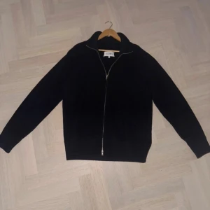 Svart kofta från Maison Margiela - Riktigt snygg marinblå kofta från Maison Margiela med hel dragkedja framtill och ribbad struktur. Tillverkad i Italien och gjord i mjuk ull. Klassisk krage och långa ärmar, perfekt för lager-på-lager under kyliga dagar. Otroligt skön! 