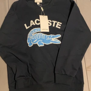 Mörkblå sweatshirt från Lacoste - Snygg mörkblå sweatshirt från Lacoste med stor blå krokodil och vit Lacoste-logga på bröstet. Tröjan har rund hals, ribbade muddar och är i regular fit. Perfekt för dig som gillar klassisk streetstyle och vill sticka ut med en ikonisk look.